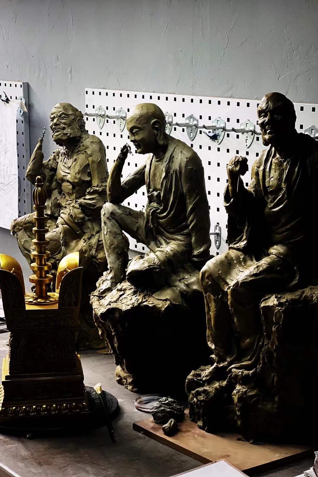 Three bronze-and-clay monk sculptures in a workshop with pegboard wall