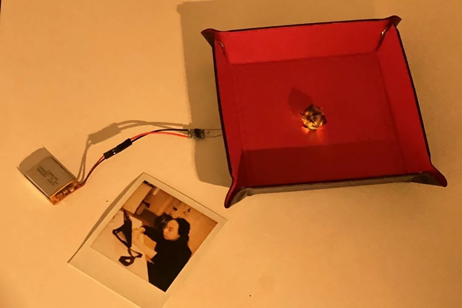 LiPo battery and circuit board feeding a glowing die in a red tray, next to a Polaroid